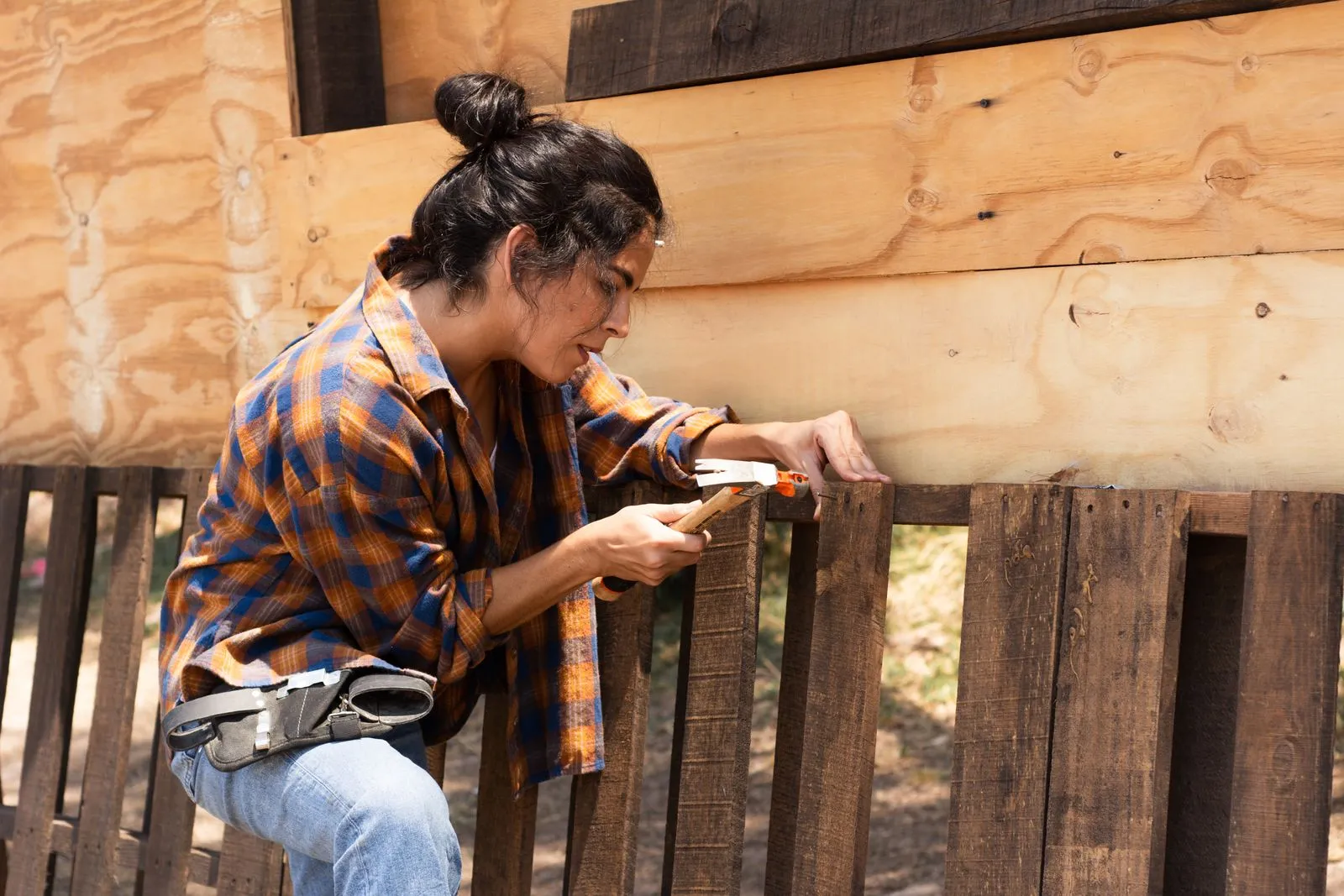 A young woman in a checked shirt and tool belt hammering a nail into a wooden fence on a sunny day